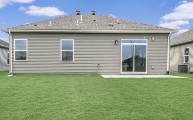 Rear view of a single-story home featuring tan vinyl siding, two large windows, a sliding glass door, and a concrete patio opening to a spacious backyard with lush green grass.