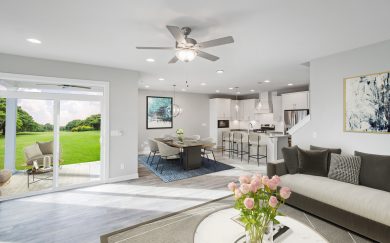 Family Room | Dining Area | Kitchen w/ Deluxe Features | Covered Patio Open-concept living area in the Emory Craftsman floor plan by Jagoe Homes, featuring a spacious family room, dining area, and upgraded kitchen with pendant lighting, white cabinetry, and a large island. Sliding glass doors provide access to the covered patio with scenic backyard views