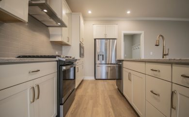 Close-up view of a modern kitchen featuring white shaker style cabinets, stainless steel appliances, Revwood flooring, and a subway tile backsplash.