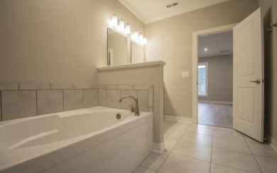 Owner&rsquo;s bathroom featuring a large soaking tub with tile surround, dual vanity lighting, and a view into the connected bedroom.