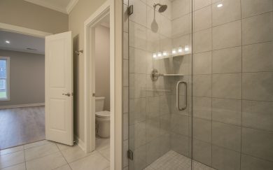 Tiled walk-in shower with glass door in owner's bathroom, next to private water closet and entrance to the bedroom.