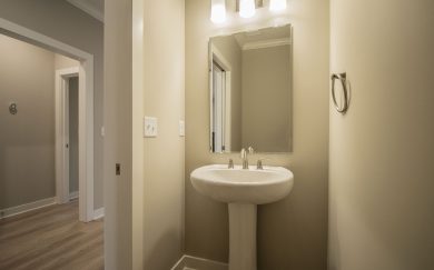 A small powder room with a pedestal sink, brushed nickel fixtures, and a rectangular mirror beneath a three-light vanity fixture.