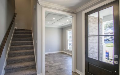 Entryway of a modern home featuring a staircase with carpeted steps to the left, a front-facing room with coffered ceiling straight ahead, and a dark front door with glass panels to the right.