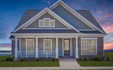 Front exterior of a charming neoclassic traditional style home at sunset, featuring a large front porch with white columns and railing, gray horizontal siding with brick accents, and a dark metal roof over the porch.