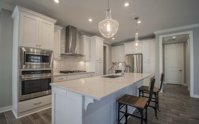 Modern open-concept kitchen featuring white shaker cabinets, stainless steel appliances, a subway tile backsplash, and a large quartz island with farmhouse-style corbels and industrial wood-and-metal bar stools.