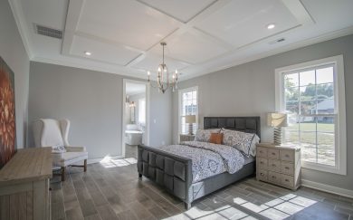 Bright and spacious primary bedroom with coffered ceiling, large windows, a tufted upholstered bed, and an en suite bathroom visible through an open doorway.