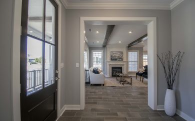Interior view from the front door of a modern home, looking into a living room with a fireplace, wood-look tile flooring, gray walls, exposed ceiling beams, and neutral-toned furniture.