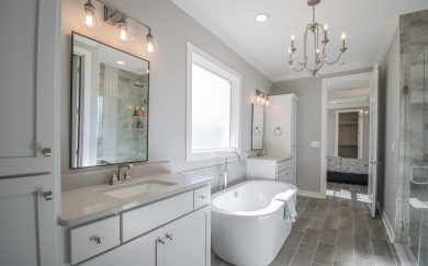 Chic primary bathroom with a freestanding tub beneath a large frosted window, dual vanities, a glass-enclosed shower, and a view into the bedroom.