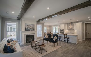 Spacious open-concept living area featuring a cozy fireplace with white trim, exposed ceiling beams, a modern kitchen with white cabinetry and pendant lighting, and a dining area with natural light from large windows.
