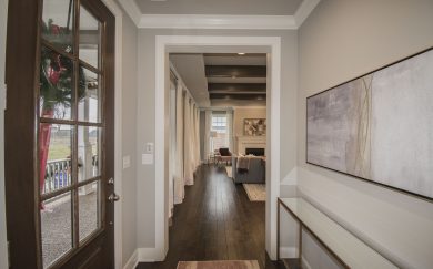 Foyer | Great Room Elegant entryway of the Merlot floor plan, showcasing a glass-paneled front door, hardwood flooring, and a view into the living room with exposed ceiling beams and a cozy fireplace. A modern console table and abstract wall art add a stylish touch.