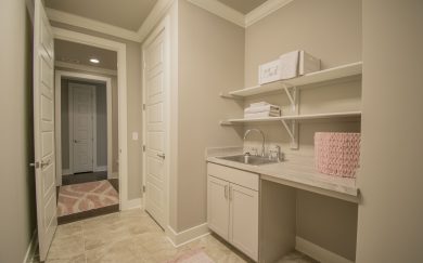 Laundry Room Functional laundry room in the Merlot floor plan featuring a built-in utility sink, ample countertop space, and open shelving for storage. Neutral tones and soft pink accents create a warm and organized environment.