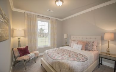 Secondary Bedroom Cozy secondary bedroom in the Merlot floor plan featuring a tufted upholstered bed with patterned bedding and accent pillows. A comfortable reading nook with a fur throw and decorative pillow is nestled beside a large window, with elegant curtains and warm lighting completing the space.