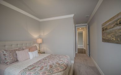 Secondary Bedroom Secondary bedroom in the Merlot floor plan featuring a tufted upholstered bed with patterned bedding and decorative pillows. The room has neutral tones, a soft carpet, and a warm table lamp. A hallway with hardwood flooring leads to additional living spaces.