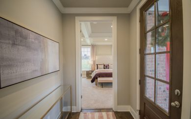 Foyer | Owner's Suite Elegant entryway of the Merlot floor plan leading to the primary bedroom. The space features a glass-paneled front door, hardwood flooring, and a modern console table with abstract wall art. The primary bedroom is visible with a tufted headboard, cozy bedding, and large windows allowing for natural light.
