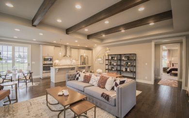Great Room | Kitchen Open-concept living area in the Merlot floor plan, featuring a cozy great room with a tufted sofa and accent pillows, a modern kitchen with a large island and pendant lights, and a dining area with natural light from sliding glass doors. Exposed ceiling beams and a view into the primary bedroom add warmth and character.