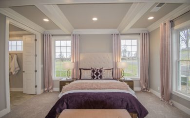 Owner's Suite Luxurious primary bedroom in the Merlot floor plan, featuring a tufted headboard, rich purple bedding, and soft pink curtains. The space is framed by large windows allowing for natural light and highlighted by elegant ceiling details. An en-suite bathroom and walk-in closet are visible through an open door.