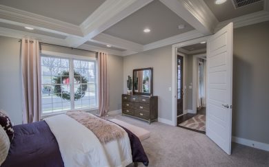 Owner's Suite Primary bedroom in the Merlot floor plan featuring a coffered ceiling, large window with pink curtains, and a view of the holiday wreath outside. The space includes a tufted bed with plush bedding, a bench, and a dark wood dresser with a mirror. The open door leads to the entryway and living area.