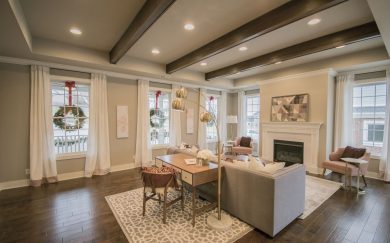 Great Room Spacious living room in the Merlot floor plan, featuring a cozy seating area with a fireplace, exposed ceiling beams, and large windows adorned with holiday wreaths. A stylish desk with a mid-century chair and an arched floor lamp adds functionality and charm.