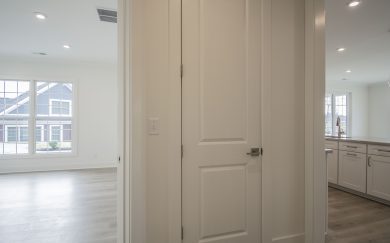 Hall to Owner's Suite Interior hallway with a storage closet, natural light flowing in from the adjacent rooms, and a view into the kitchen with white cabinetry and Revwood floors.