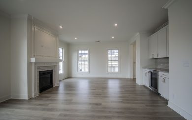 Great Room | Beverage Center Wide-angle view of the Malbec’s open-concept great room featuring a fireplace with decorative trim, large windows providing natural light, and a built-in beverage station with white cabinetry.
