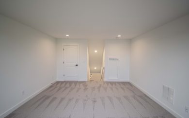 Interior view of the upstairs bonus room in the Emory Craftsman, showing freshly carpeted flooring, led lighting, a door to a storage closet, and an open stairwell leading to the main level.