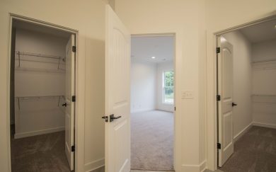 View from the owner&rsquo;s bathroom showing two separate walk-in closets with wire shelving and a doorway leading into the carpeted bedroom with natural light from large windows.