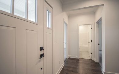 Interior view of the Emory Craftsman home&rsquo;s front entry, featuring a white door with glass inserts, revwood flooring, and a hallway leading to bedrooms and a bathroom.