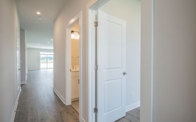 View from the hallway in the Leonardo Craftsman home, showcasing a bathroom and bedroom entry, leading to the open-concept living area with natural light streaming in.