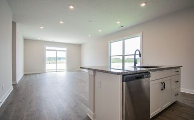 Open-concept living and dining area in the Leonardo Craftsman, viewed from the kitchen island with stainless steel dishwasher, granite countertops, and large windows offering natural light and views of the outdoors.