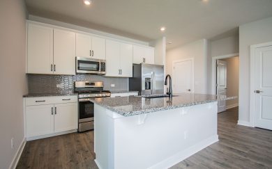 Modern kitchen in the Leonardo Craftsman featuring white cabinetry, granite countertops, stainless steel appliances, a center island with an undermount sink, and a textured gray tile backsplash.