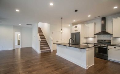 Open-concept kitchen and living area in the Emory Craftsman featuring revwood flooring, a large island with black countertops, white cabinets, stainless steel appliances, and a staircase leading to the bonus room