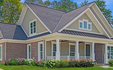 Side view of the Moscato floor plan with taupe siding, brick accents, a deep front porch with white columns, and colorful blooming flower beds lining the foundation.