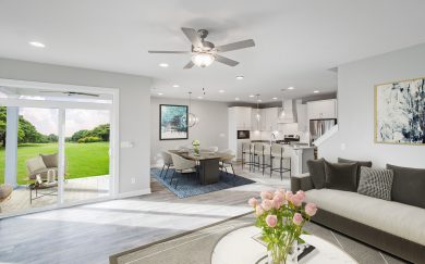 Beautifully staged open-concept living area with a deluxe kitchen, dining area, and family room; featuring white cabinetry, pendant lighting, revwood floors, and sliding glass doors opening to a covered patio and lush green backyard.