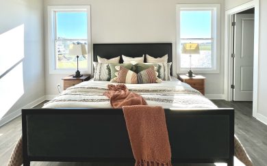 Front-facing view of a stylishly furnished owner&rsquo;s suite featuring a dark upholstered bed frame, layered pillows, wood nightstands, and two large windows with lake views.