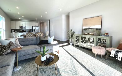 Spacious and sun-filled living room in the Leonardo Craftsman home, featuring a large sectional sofa, accent tables, a green mirrored sideboard, and open views to the kitchen with white cabinetry and stainless steel appliances.