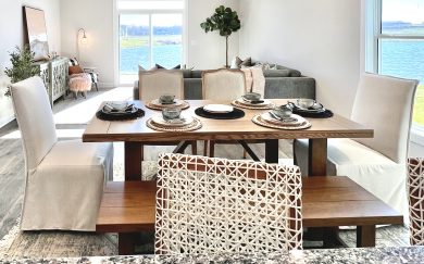 Bright and inviting dining and living space in the Leonardo Craftsman home, featuring a wooden table set for six, cozy upholstered chairs and benches, and panoramic lake views through sliding glass doors and large windows.