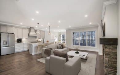 Open Concept Living | Deluxe Kitchen Open-concept living space featuring a modern kitchen with white cabinetry and stainless steel appliances, a cozy living room with a stone fireplace and mounted TV, and a dining area with large windows offering wooded views.
