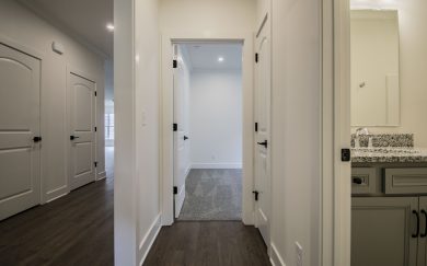 Optional Bedroom 3 in lieu of Flex Space View of a hallway leading to a carpeted bedroom, with Revwood flooring, a bathroom vanity with granite countertop to the right, and multiple white paneled doors.