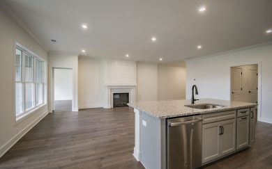 Great Room with Electric Fireplace Open-concept living area with a large granite island, stainless steel dishwasher, wide windows, Revwood floors, and an electric fireplace with a white wood mantel.